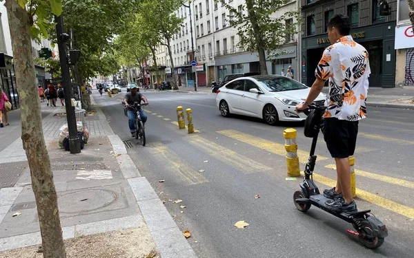 Un homme en chemise fleurie conduit une trottinette électrique dans une rue de Paris, avec un cycliste et une voiture blanche en arrière-plan
