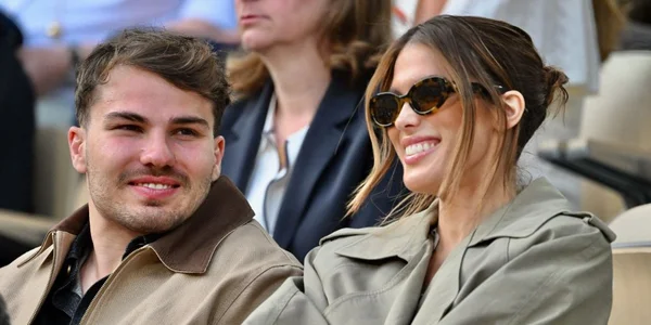 Antoine Dupont et Iris Mittenaere assis côte à côte en extérieur, lui en veste beige à col marron, elle en lunettes de soleil et trench-coat vert clair, lors d'un événement public.