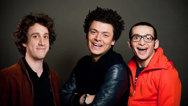 Portrait des trois acteurs principaux de la série Soda, souriant devant un fond gris foncé, avec l'un portant une veste marron, un autre un haut à manches en cuir noir et un troisième un sweat orange à capuche et des lunettes.