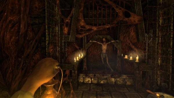 Capture d'écran de gameplay d'Amnesia: The Dark Descent montrant une main tenant une lanterne dans une pièce sombre aux murs de pierre, avec une figure squelettique enchaînée à une structure en bois éclairée par des bougies.