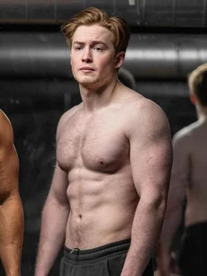 Photographie de l'acteur Kit Connor, torse nu, avec des cheveux roux-blonds et une carrure musclée, posant dans un intérieur évoquant une salle de sport, regardant légèrement hors cadre.