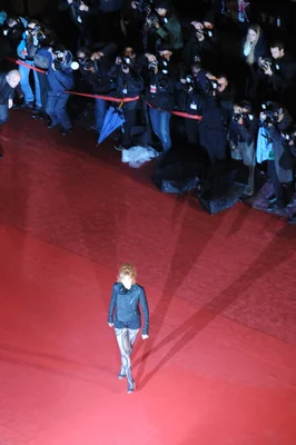 Mylène Farmer, aux cheveux blonds bouclés, marchant sur un tapis rouge vêtue d'une veste noire et de collants à motifs, entourée de photographes derrière une barrière.