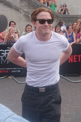Kit Connor, vu de face, portant des lunettes de soleil, un t-shirt blanc et un pantalon sombre, lors de la première de The Gray Man à Londres, avec des barrières promotionnelles du film en arrière-plan.