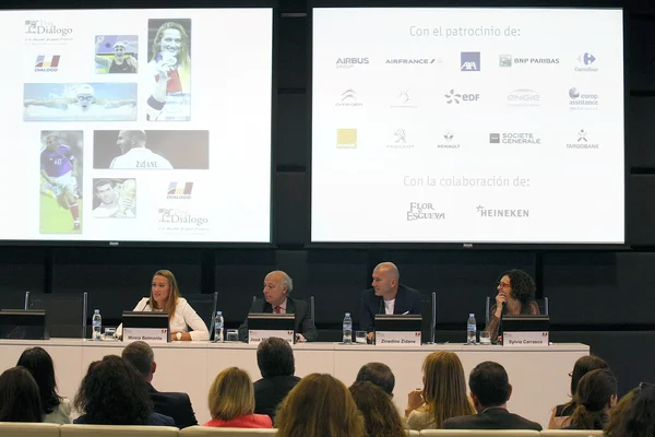 Zinedine Zidane lors d'une table ronde au XII Prix Diálogo, assis à une longue table avec des plaques nominatives, devant de grands écrans de projection affichant des logos de sponsors et des images d'athlètes.