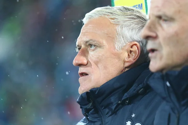 Didier Deschamps, entraîneur de l'équipe de France, en plan rapproché de profil, les cheveux blancs, vêtu d'une veste bleu foncé, regardant intensément vers la gauche.