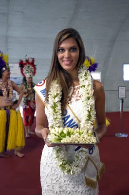 Iris Mittenaere, Miss France 2016, en robe blanche en dentelle et écharpe, tenant un plateau de fleurs blanches sur un tapis rouge, entourée de deux personnes en costumes polynésiens colorés.