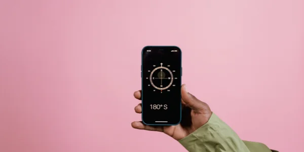 Main tenant un smartphone affichant une application de boussole indiquant '180° S' sur un fond rose, illustrant l'utilisation des boussoles numériques sur mobile.