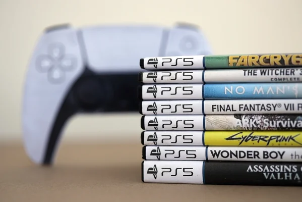 Pile de boîtiers de jeux PlayStation 5 au premier plan, avec une manette DualSense blanche floue en arrière-plan, illustrant la transition vers le numérique.