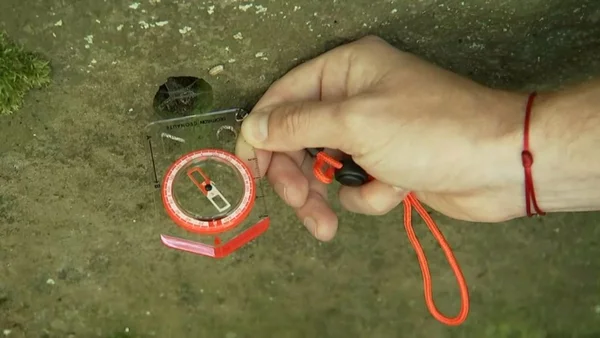 Gros plan d'une main tenant une boussole à base transparente avec un cadran orange et un cordon, posée sur une surface en béton moussu, illustrant les relevés magnétiques dans le village de Laschamps.