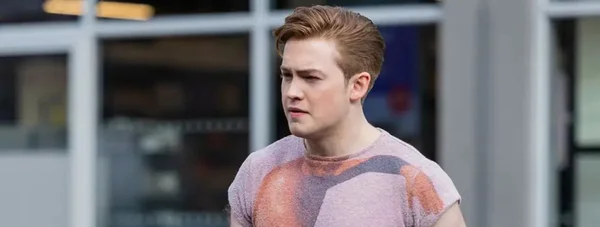 Photographie en plan moyen de l'acteur Kit Connor, cheveux bruns courts, portant un t-shirt à motifs, debout à l'extérieur devant une façade d'immeuble floue, regardant sur le côté avec une expression neutre.