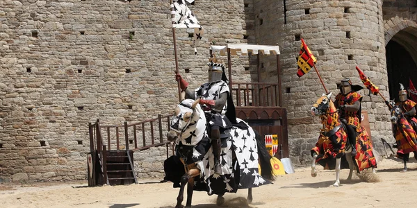 Chevaliers en armure complète et tabards héraldiques colorés montant des chevaux lors d'une reconstitution médiévale ou d'un tournoi de joutes devant un mur de château en pierre, pendant la Fête des Remparts en Bretagne.