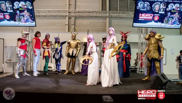 Cosplayers costumés en personnages de Saint Seiya sur la scène du Hero Festival à Marseille, avec de grands écrans affichant des informations sur l'événement en arrière-plan.
