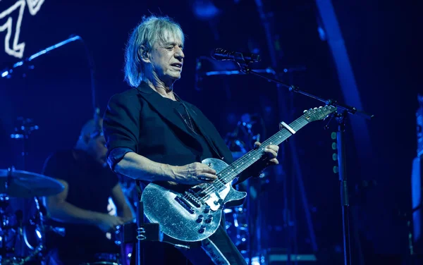 Jean-Louis Aubert, cheveux blonds mi-longs, jouant de la guitare électrique argentée et chantant sur scène sous un éclairage bleu, avec un batteur en arrière-plan flou.