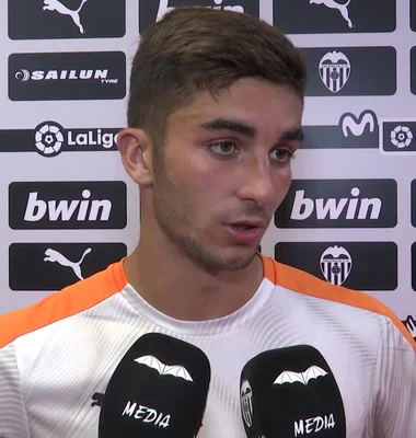 Ferran Torres, attaquant espagnol, interviewé par la télévision du Valencia CF en 2019, portant un maillot d'entraînement blanc et orange, souriant devant des micros et des logos de sponsors.