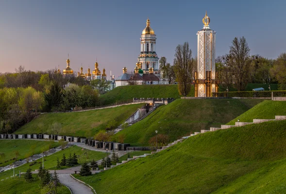 Vue panoramique du complexe de la laure de Kiev-Petchersk au crépuscule, avec ses églises aux dômes dorés et son clocher blanc, entourés de pentes herbeuses en terrasses.