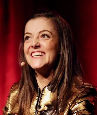 Camille Lellouche souriante sur scène, veste en sequins dorés et micro-casque, fond rouge, captant l'attention du public.
