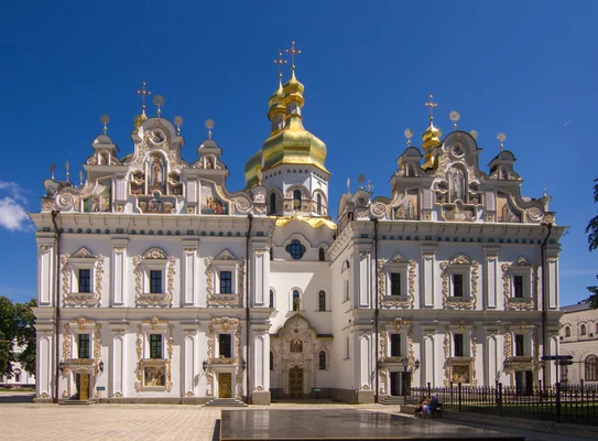 L'église de la Trinité de la laure des Grottes de Kiev, murs blancs ornés de décorations baroques dorées, surmontés de dômes en bulbe dorés sous un ciel bleu clair.