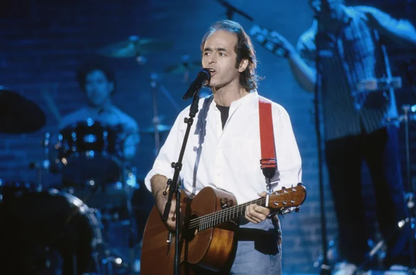 Jean-Jacques Goldman, cheveux bruns et chemise blanche, chantant dans un micro en jouant de la guitare acoustique sous un éclairage bleu