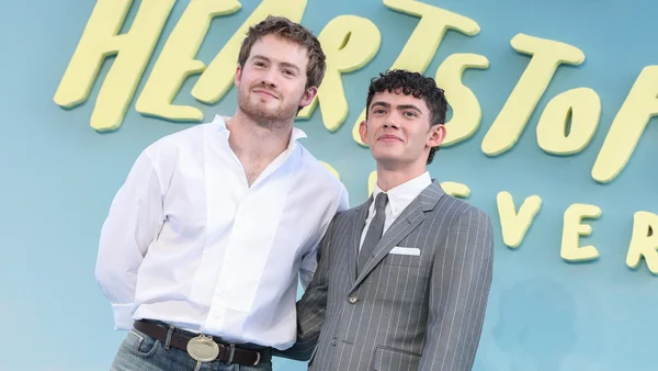 Kit Connor et Joe Locke posent lors de l'avant-première de Heartstopper Forever à Londres, devant un mur bleu clair avec le titre de la série en lettres jaunes. Kit porte une chemise blanche et un jean, Joe un costume gris rayé.