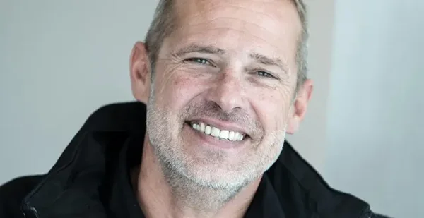 Portrait de l'écrivain Fabien Clauw, souriant, les cheveux grisonnants courts et la barbe naissante, vêtu d'une veste noire, devant un fond clair.