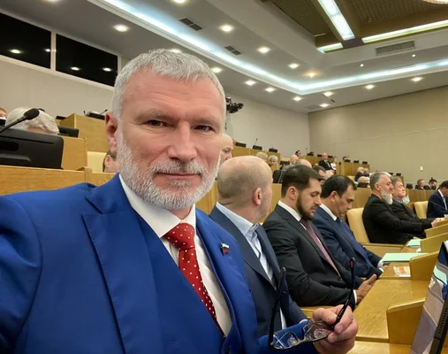 Alexeï Jouravlev, député de la Douma d'État de la Fédération de Russie, prenant un selfie dans une salle d'assemblée, en costume bleu et cravate rouge.