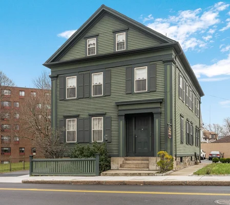 La maison Lizzie Borden à Fall River, une maison en bois vert à deux étages avec des volets sombres et un toit à pignon, située à un coin de rue sous un ciel bleu, avec un immeuble en briques en arrière-plan.