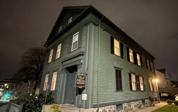 Photographie nocturne de la maison Lizzie Borden à Fall River, un bâtiment en bois vert à deux étages avec des fenêtres éclairées et une enseigne suspendue indiquant 'The Mystic Garden'.