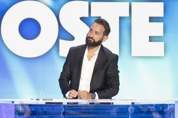 Cyril Hanouna, barbu, en veste noire et chemise blanche, assis à son bureau devant un fond bleu avec les lettres blanches 'OSTE', lors de l'émission TPMP sur W9.