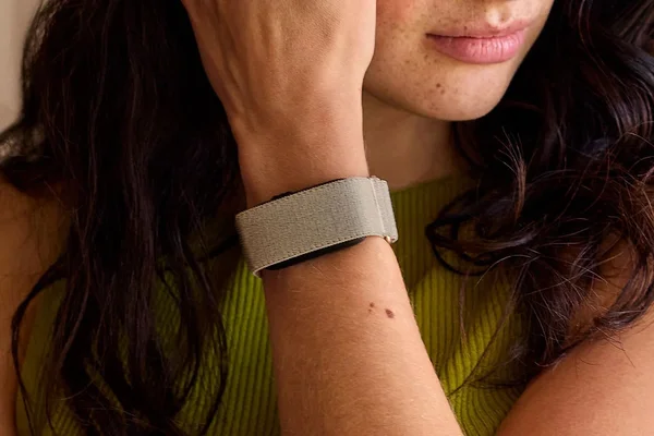 Plan rapproché d'une personne aux cheveux bruns et taches de rousseur portant un bracelet en tissu gris clair au poignet gauche, le menton appuyé sur la main, vêtue d'un haut côtelé vert