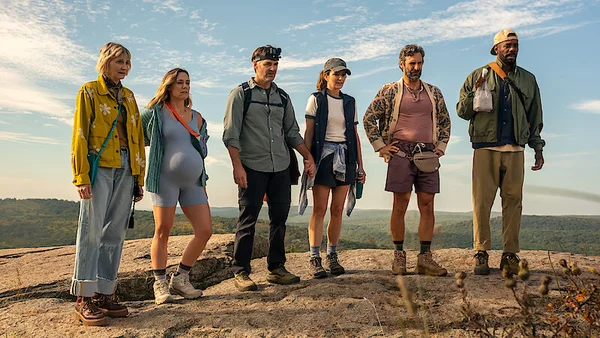 Groupe de six personnes, dont une femme enceinte et un homme portant une lampe frontale, alignés au bord d'une falaise rocheuse surplombant un paysage forestier sous un ciel bleu, extrait promotionnel de la série The Four Seasons sur Netflix.