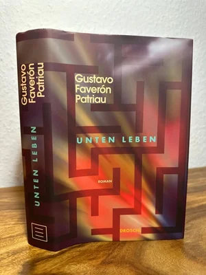 Photographie du livre « Unten leben » de Gustavo Faverón Patriau, posé debout sur une surface en bois contre un mur blanc, illustrant l'édition allemande de « Les lieux souterrains ».
