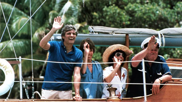 Quatre personnes, dont un homme en polo bleu et une femme portant un chapeau de paille, debout sur le pont d'un voilier, saluant la caméra, avec une végétation verdoyante en arrière-plan, extrait du film The Four Seasons de 1981.