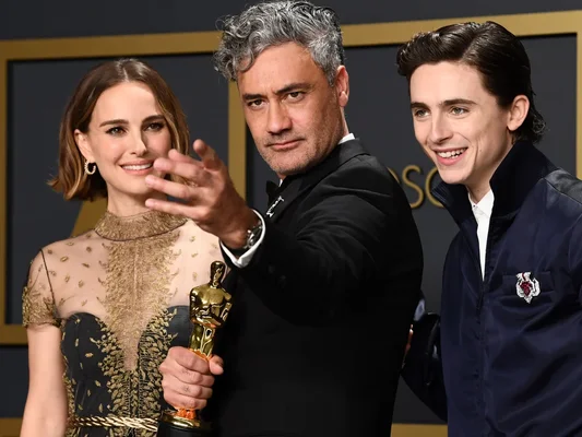 Taika Waititi, réalisateur choisi pour le prequel de Charlie et la Chocolaterie, tenant sa statuette des Oscars lors d'une cérémonie, entouré de deux personnes en tenue de soirée.