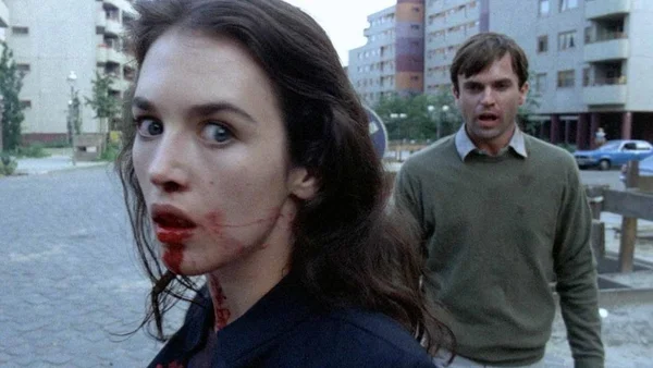 Isabelle Adjani couverte de sang dans une scène du film Possession d'Andrzej Zulawski, regardant en arrière avec un air choqué, tandis qu'un homme se tient derrière elle dans un décor urbain