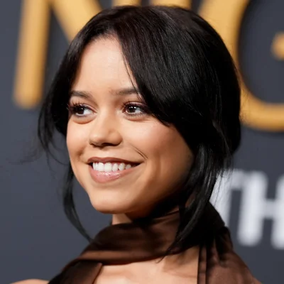 Jenna Ortega souriante, les cheveux noirs cerise avec frange, portant un haut marron, devant un fond flou avec de grandes lettres dorées