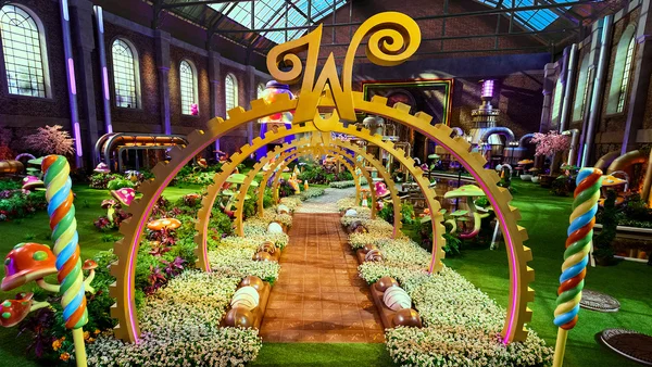 Installation décorative sur le thème de Willy Wonka : arche dorée géante, sucettes géantes et champignons colorés dans une salle en briques avec fenêtres en arc.