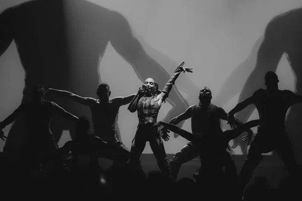 Photographie en noir et blanc d'un concert de FKA Twigs à l'Adidas Arena, où l'artiste, micro en main, bras levé, est entourée de danseurs en silhouette, éclairés par un grand écran diffusant des ombres géantes.
