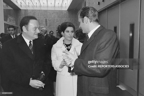 Photographie en noir et blanc de René Goscinny et Albert Uderzo lors de la première du film « Astérix », discutant avec une femme ; Goscinny tient une cigarette, Uderzo fait un geste.