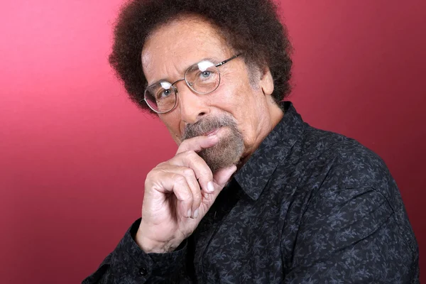 Portrait de Jean Schultheis en octobre 1983, arborant une coupe afro, des lunettes et une barbiche, vêtu d'une chemise noire à motifs, la main sous le menton, devant un fond rouge.