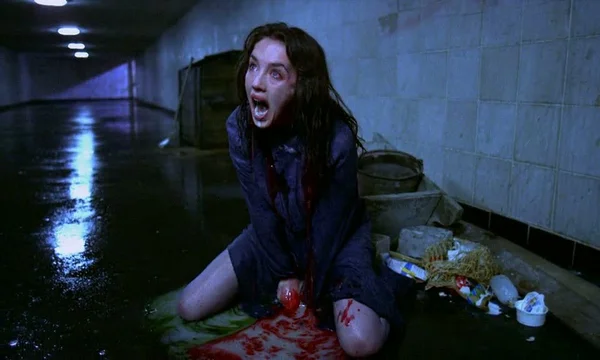 Scène du film Possession d'Andrzej Żuławski : une femme aux cheveux mouillés et au chemisier taché de sang, agenouillée dans un couloir inondé, hurlant la bouche grande ouverte, sous une lumière bleue froide