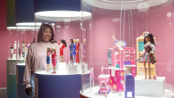 Femme aux cheveux bruns portant un sweat rose souriant devant des vitrines cylindriques exposant des poupées Barbie et des accessoires, dans un musée aux murs roses.