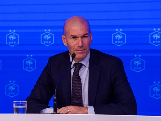Zinédine Zidane en costume sombre, lors d'une conférence de presse de l'équipe de France, assis à une table avec un micro et un verre d'eau, devant un fond bleu orné du logo de la Fédération Française de Football.