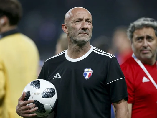 Fabien Barthez, chauve et barbu, vêtu d'un maillot noir Adidas avec l'écusson 'France 98', tenant un ballon de football blanc à motifs pixellisés, sur un terrain de sport, regardant au loin.