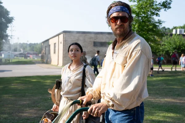 Un homme barbu portant lunettes de soleil et bandana bleu pousse un chariot aux côtés d'une femme en tenue beige sur un champ herbeux, près d'un bâtiment en briques, sous une lumière vive.