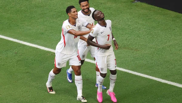 Trois joueurs de l'équipe d'Angleterre en maillot blanc célèbrent sur la pelouse verte, le joueur au premier plan portant des crampons roses, regardant vers le ciel, entouré de ses coéquipiers.