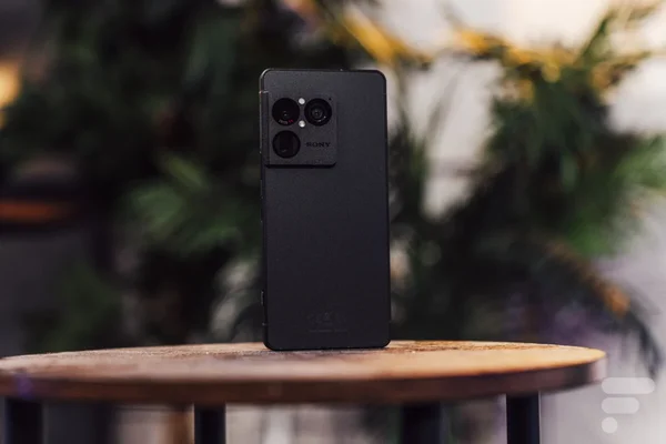 Smartphone Sony Xperia 1 VIII noir posé verticalement sur une table en bois ronde, montrant son module caméra arrière et sa finition texturée, avec un arrière-plan flou de plantes d'intérieur.