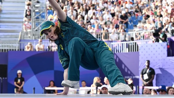 Rachael Gunn, connue sous le nom de Raygun, en tenue vert et jaune, exécute une figure de breakdance sur une main lors des Jeux olympiques de Paris 2024, devant une foule nombreuse.