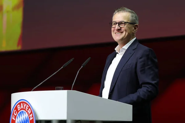 Jan-Christian Dreesen, président-directeur général du FC Bayern München, souriant derrière un pupitre blanc au logo du club lors d'une assemblée générale annuelle.