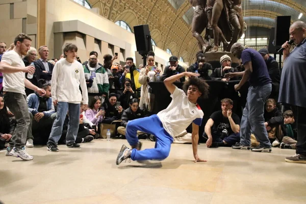 Un breakdancer de l'équipe de France de breakdance, vêtu d'un t-shirt blanc et d'un pantalon de survêtement bleu, exécute une figure au sol au centre du Musée d'Orsay, entouré d'un public nombreux, sous les voûtes majestueuses du musée.