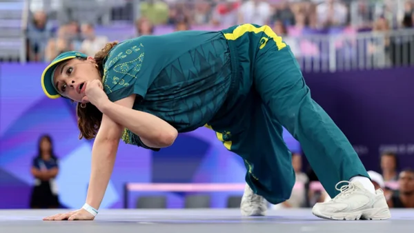 La B-Girl Raygun de l'équipe d'Australie en survêtement bleu sarcelle et jaune avec casquette assortie, exécutant une figure de breakdance au sol lors des épreuves de breakdance aux Jeux Olympiques de Paris 2024, à la Place de la Concorde, devant un public flou et des éléments de branding violets.